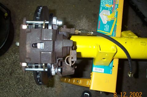 1986 Ford Bronco - Overhead close-up of a rear disc brake conversion mounted on a yellow axle housing: rusty cast caliper with slide pins, coiled parking-brak…
