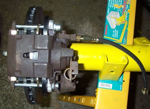 1986 Ford Bronco - Top-down view of a rear disc brake assembly on a yellow axle housing, showing the caliper bridge bolts, vented rotor edge, parking-brake sp…