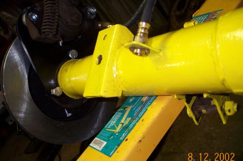 1986 Ford Bronco - Detail of a yellow-painted axle tube where a welded spring perch and shock-mount bracket meet the housing end, with a brass fitting and rub…