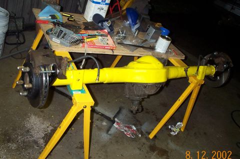 1986 Ford Bronco - Full-length view of a yellow-painted rear axle housing on two yellow sawhorses, with rusty disc brake calipers and rotors hung on both ends…