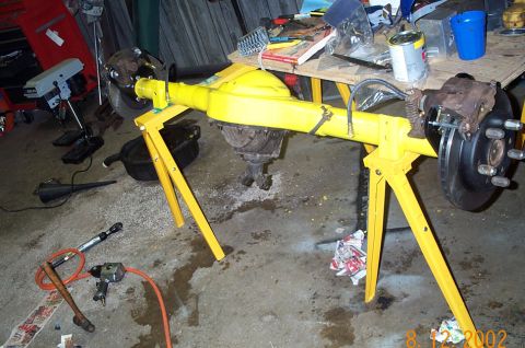 1986 Ford Bronco - A freshly yellow-painted solid rear axle assembly sitting across two yellow sawhorse stands in a garage, with a disc brake rotor and five-l…