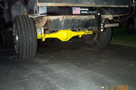 1986 Ford Bronco - Night shot from behind a black truck on a driveway, looking under the rusty rear bumper at a bright yellow solid axle spanning between whit…