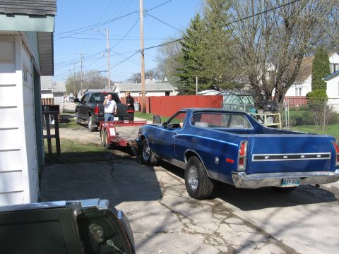 1976 Ford Ranchero - A blue Ford Ranchero is being towed on a trailer by a black pickup truck on a sunny day.