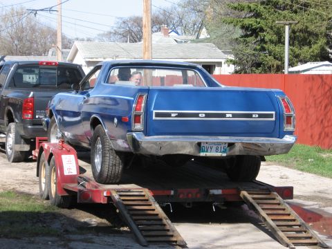 1976 Ford Ranchero - A blue 1976 Ford Ranchero is loaded onto a trailer, being towed by a black pickup truck.