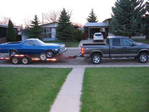 1976 Ford Ranchero - Pickup truck tows a blue 1976 Ford Ranchero on a trailer down a residential street.