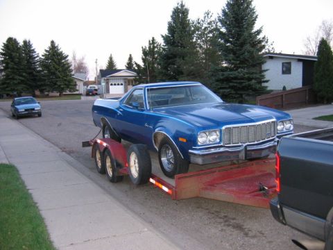 1976 Ford Ranchero - A blue 1976 Ford Ranchero is loaded onto a red trailer, towed by a dark pickup truck on a suburban street.