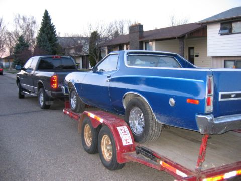 1976 Ford Ranchero - A blue coupe utility is loaded onto a trailer, towed by a pickup truck on a residential street.