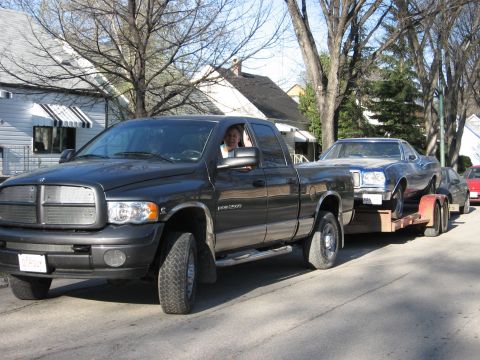 1976 Ford Ranchero - A dark-colored Dodge Ram pickup truck tows a blue car on a trailer, with a person visible in the driver's seat.
