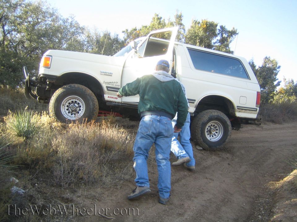 White SUV with off-road tires on a dirt path, driver's door open, two men standing nearby