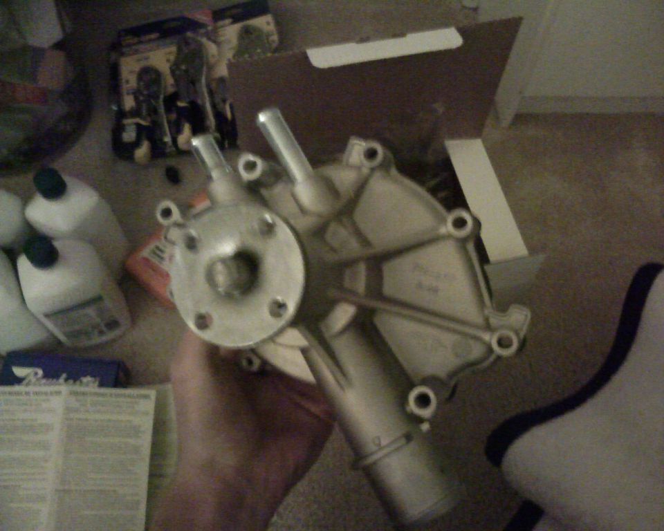 Hand holding a metal water pump with two spouts and a circular mounting flange. Other car parts and bottles are visible in the background