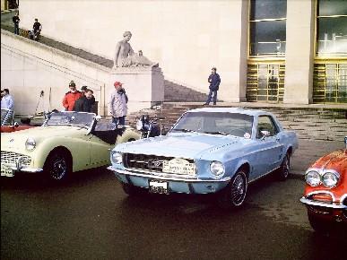 1991 Ford Crown Victoria - A light blue 1967 Ford Mustang coupe is parked in front of a building with a yellow convertible and an orange car nearby.