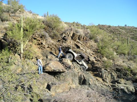 1966 Ford Bronco - An off-road vehicle is stuck on a steep, rocky incline in a desert environment, with two individuals observing the situation.