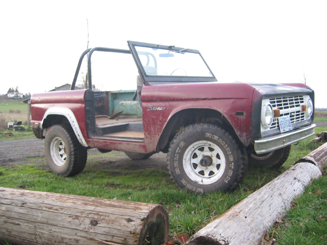 Red 1968 Ford Bronco with doors removed, parked on grass with logs in foreground