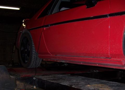 1988 Pontiac Fiero - A red 1988 Pontiac Fiero is positioned on a lift, showing its side profile with black wheels.