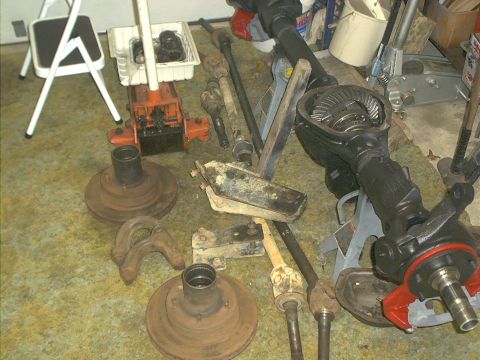1986 Ford Bronco - Various axle parts, including a differential housing and brake rotors, are laid out on the floor next to an orange hydraulic jack.
