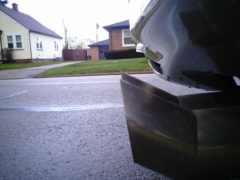 Close-up of a car's rear bumper and exhaust pipe. Houses and a street are visible in the background