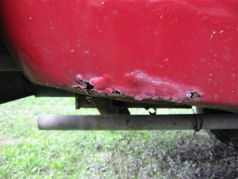 1974 Ford F-100 - Rust has severely compromised the lower body panel of this red truck, with an exhaust pipe visible below.