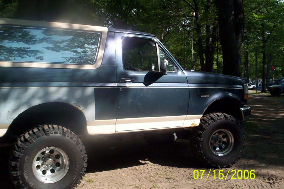 Lifted dark blue Ford Bronco with big mud tires and alloy wheels, side view in a wooded dirt area, dated 07/16/2006