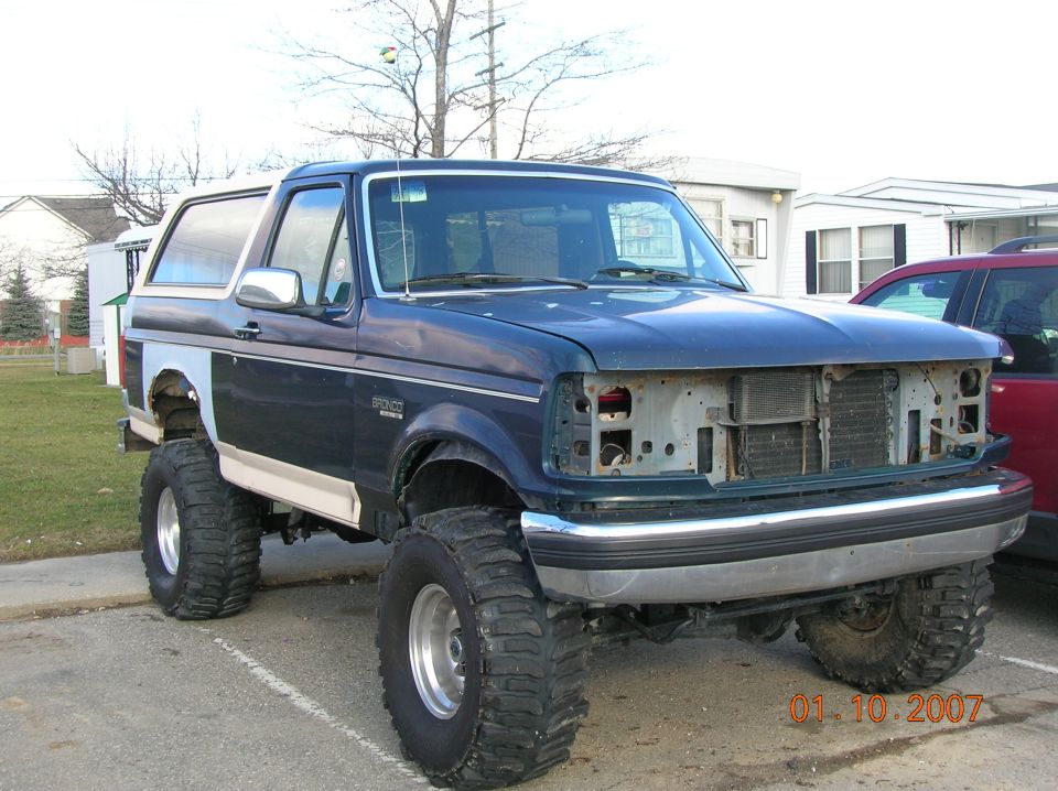 Lifted blue Ford Bronco with grille removed on big mud tires in a parking lot