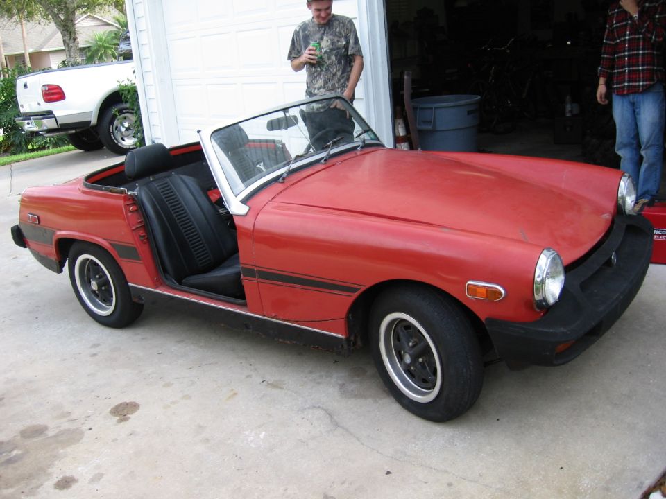 Red convertible sports car with black racing stripes and black interior parked in front of a garage. A white pickup truck is visible in the background