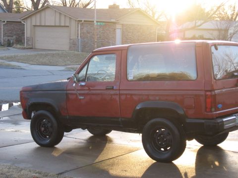 1984 Ford Bronco II - The sun glints off the side of a red 1984 Ford Bronco II, parked in front of a suburban house.