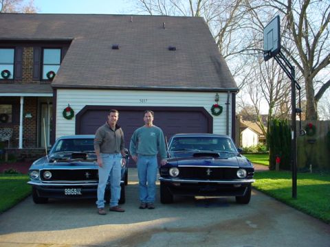 1970 Ford Mustang - Two classic Ford Mustangs are parked in front of a suburban home, one with its hood raised.