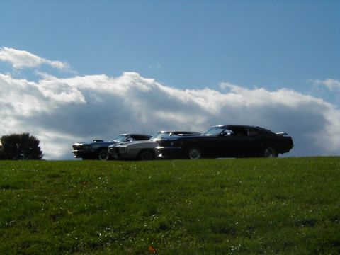 1970 Ford Mustang - Three classic cars are lined up on a grassy incline against a dramatic sky.