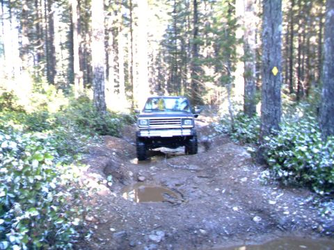 1987 Ford Bronco II - A dark blue 1987 Ford Bronco II navigates a muddy, rutted trail through a dense forest.