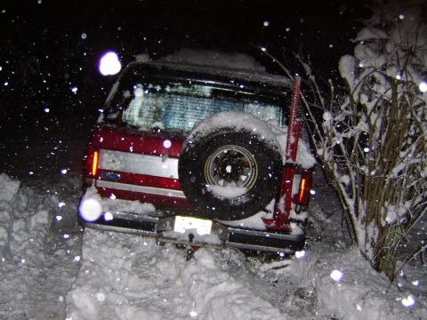 1989 Ford Bronco - A red SUV is stuck in a snowbank at night with snow falling around it.