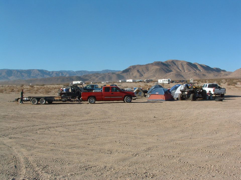 Desert campsite with red pickup truck, trailer, off-road vehicle, white pickup truck, and tents