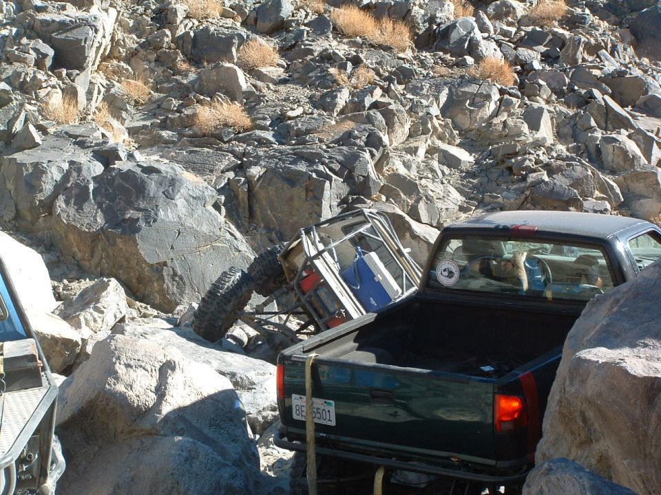 Off-road buggy tipped on its side amidst rocks, with a dark green pickup truck behind it