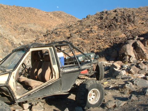 1994 Jeep YJ - A heavily modified off-road buggy and a green Jeep sit in a rocky desert terrain under a clear blue sky.