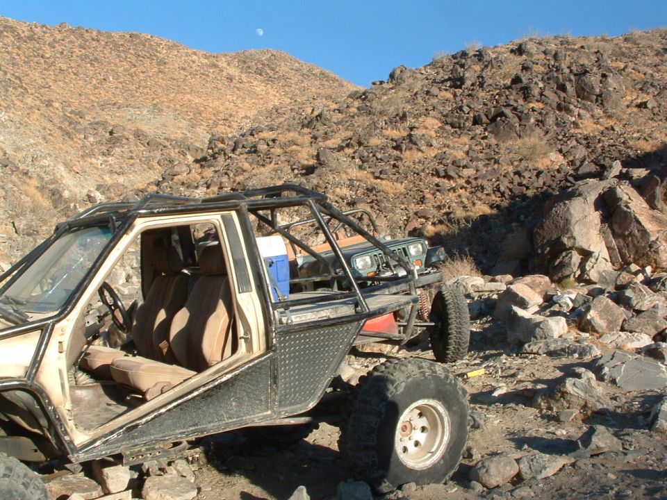 Off-road buggy with roll cage and tan interior, parked next to a green Jeep in a rocky, desert landscape