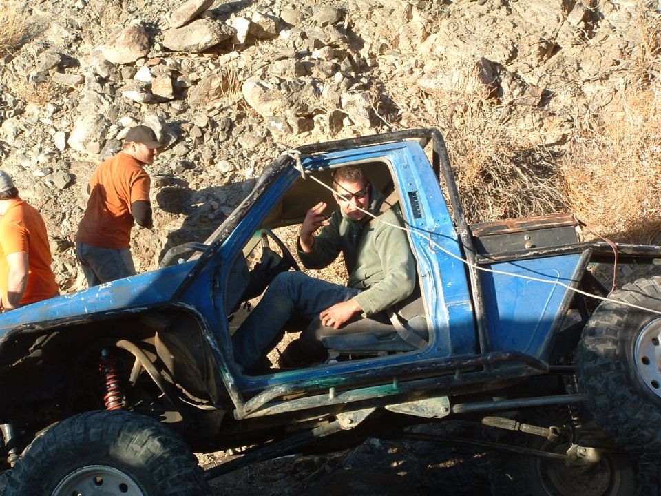 Modified blue off-road truck with people, one driver inside, others standing nearby on rocky terrain