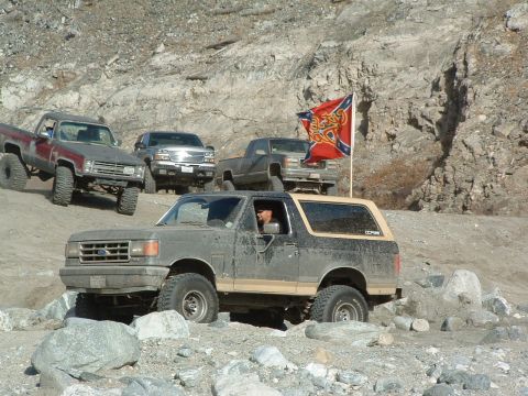 1994 Jeep YJ - Several trucks, including a Ford Bronco, navigate a rocky off-road trail. A flag is mounted on one of the trucks.