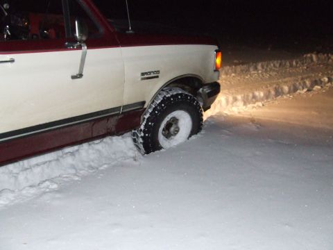 1990 Ford Bronco - A two-tone white and maroon Ford Bronco is stuck in deep snow, with its front wheel partially submerged.