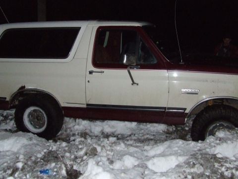 1990 Ford Bronco - A two-tone Ford Bronco is partially submerged in snow, showing its side profile and a "Bronco" emblem.