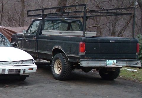 1989 Ford F-250 - A black Ford pickup truck with a ladder rack and toolbox is parked next to a white car.