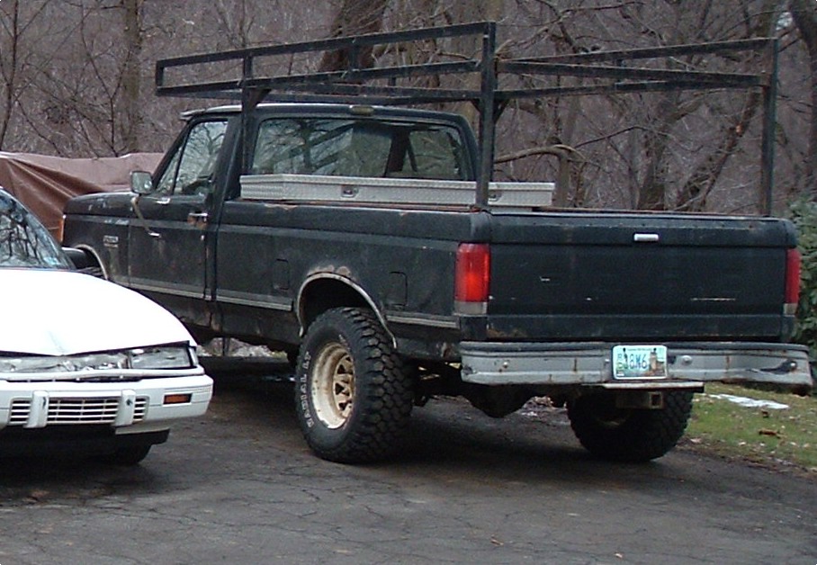 Black Ford pickup truck, rear view, with a ladder rack and toolbox. Visible rust on the body