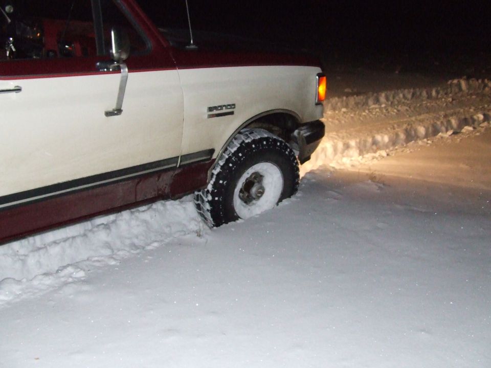 White and maroon Ford Bronco stuck in deep snow at night, front tire buried