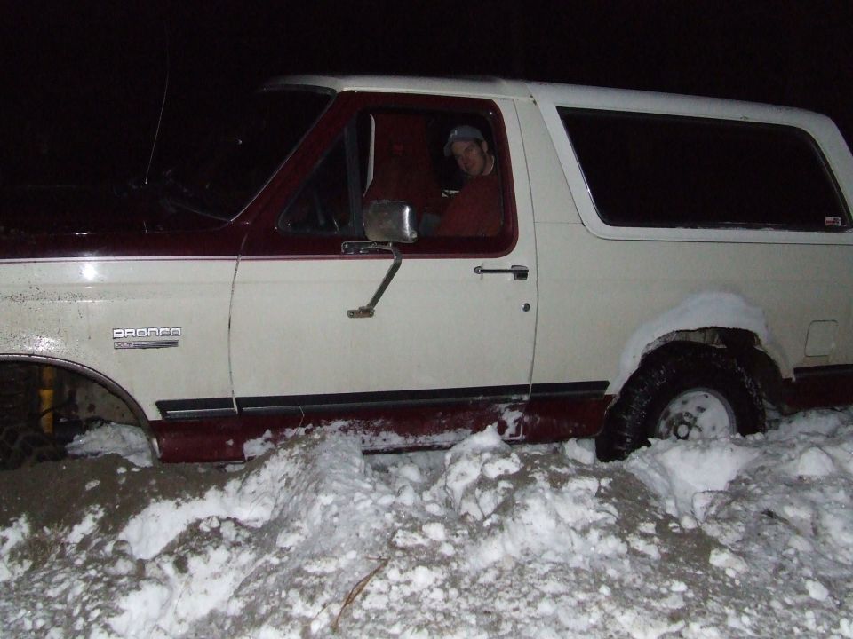 White and maroon Ford Bronco stuck in deep snow at night with driver looking out the window
