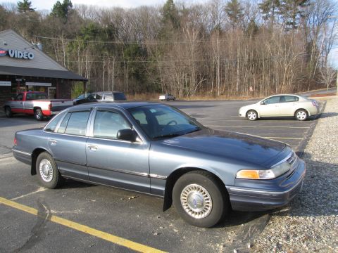 1991 Ford LTD Crown Victoria - A blue sedan is parked in a lot with other vehicles and a building in the background.