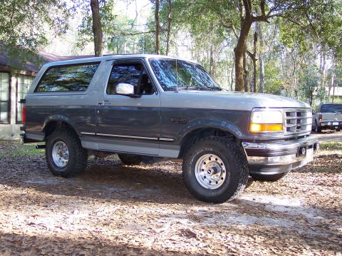 1996 Ford Bronco - A two-tone gray Ford Bronco is parked on a dirt lot, with another vehicle visible in the background.