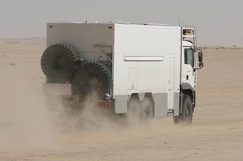 1993 Ford Bronco - A white expedition truck kicks up dust as it drives through a desert landscape, equipped with two spare tires mounted on the rear.