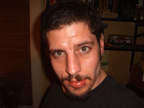 1993 Ford Bronco - Indoor close-up portrait of a person with dark curly hair, a goatee, lip and ear piercings, wearing a black t-shirt, with a shelf holding a…