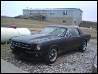 1965 Ford Mustang - A dark colored 1965 Ford Mustang coupe is parked on a gravel lot with a commercial building in the background.