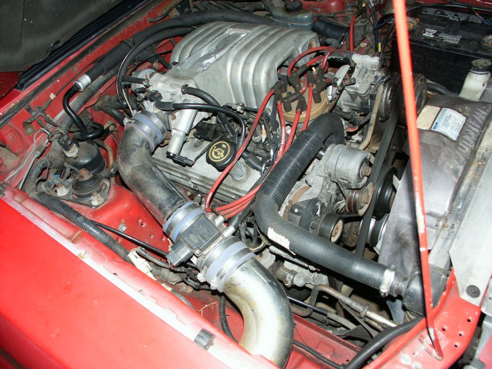 Engine bay of a 1990 Ford Mustang, showing the V8 engine, intake, and various hoses and wires