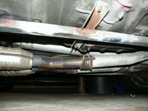 1990 Ford Mustang - The underside of a vehicle shows the exhaust system, including the catalytic converter and muffler, with visible rust on mounting brackets.