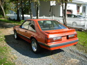 1990 Ford Mustang - This orange 1990 Ford Mustang coupe is shown from the rear, parked on a gravel driveway.