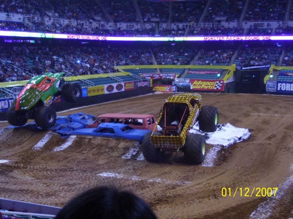 Monster trucks in an arena, one with a green and red design, the other yellow with black spots, crushing cars